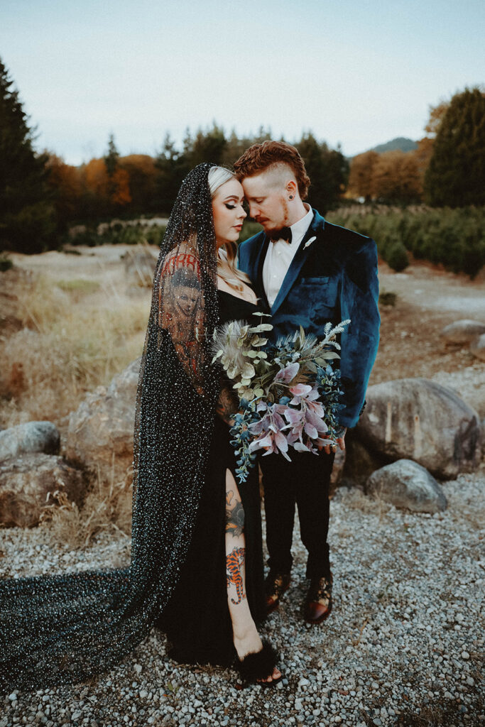 A bride dressed in an untraditional black wedding dress with black glitter veil and a groom wearing a velvet teal jacket share a moment during their First Look.