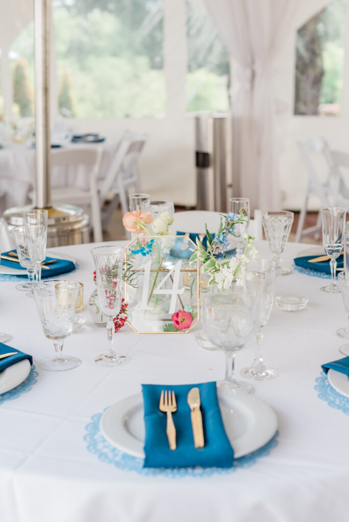 Circular guest table set with gold flatware, crystal goblets, and navy napkins. Floral bud vases and votives decorate the center.