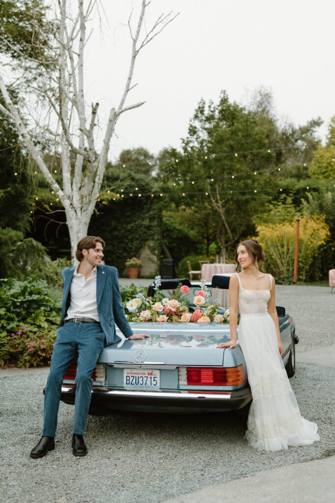 Bride and groom posing beside a vintage car in the gardens at Twin Willow Gardens, a charming Pacific Northwest garden wedding venue.