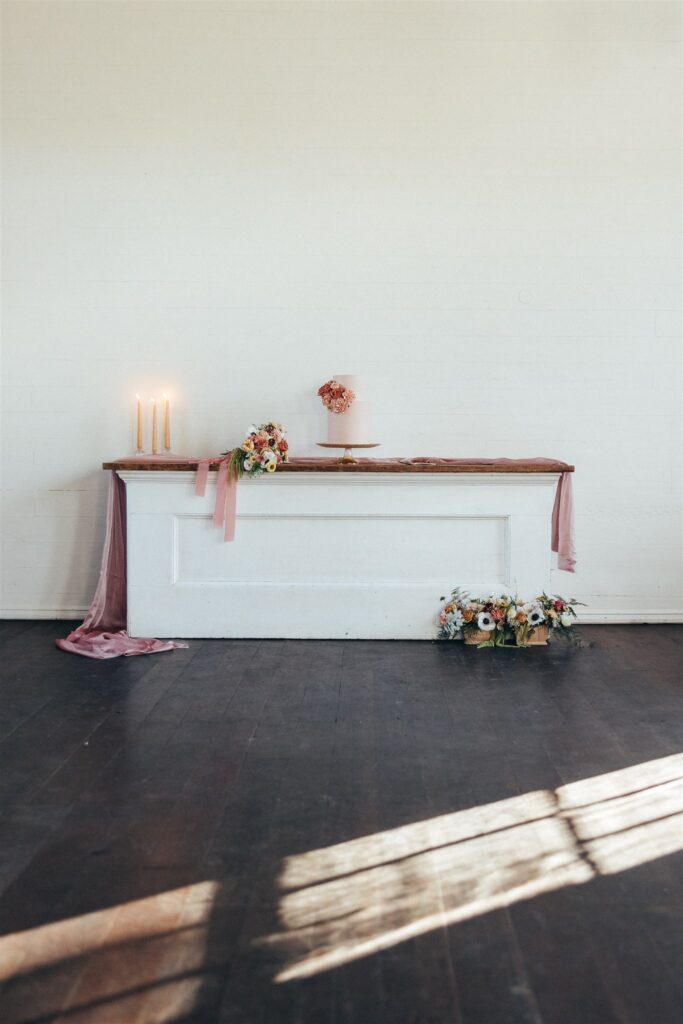 Two tier pink cake sitting on top of a white buffet accompanied by two floral arrangements in pinks, oranges, and whites.