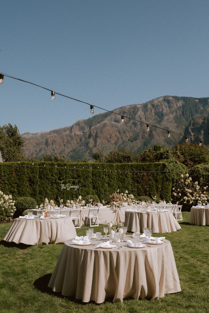 Round guest tables draped in beige linens and white napkins sitting amongst the Cape Horn Estates garden and lawn with views of the mountains in the bakcground